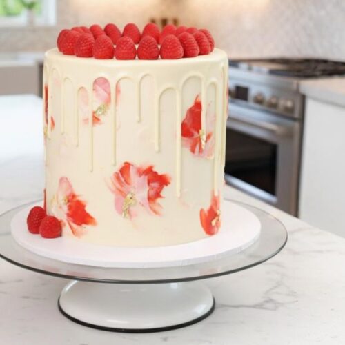 A tall white chocolate raspberry cake with pink ombre frosting, white chocolate drip, fresh raspberries and flowers on top, displayed on a white cake stand in a minimalistic kitchen.