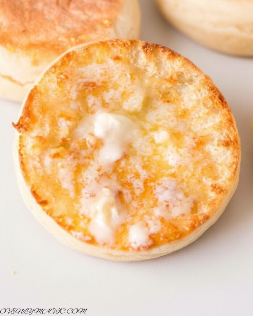 Freshly baked English Muffins split open, showing soft, airy interior (“nooks and crannies”), on a plate or wooden board.