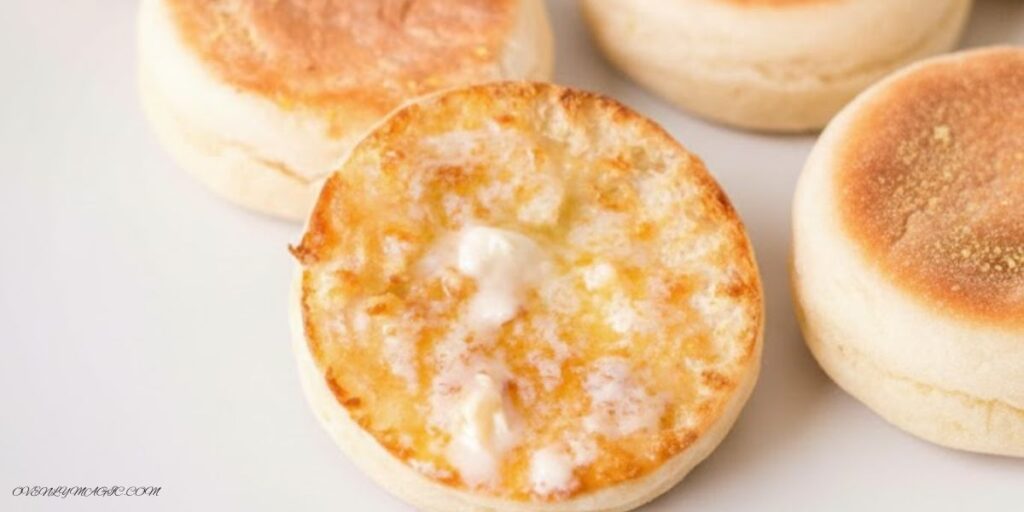 Freshly baked English Muffins split open, showing soft, airy interior (“nooks and crannies”), on a plate or wooden board.